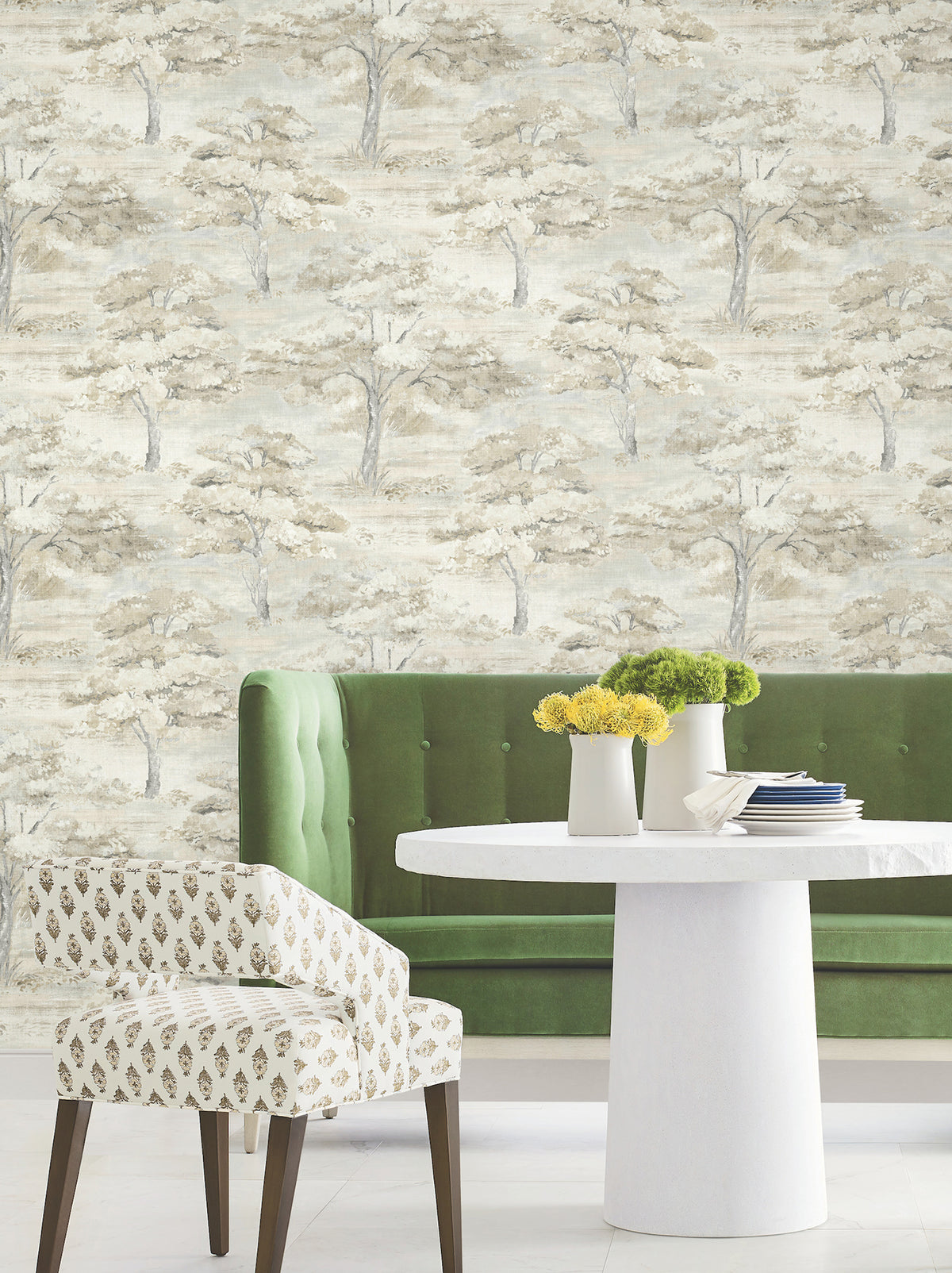 A green velvet tufted bench and a patterned chair sit by a round white table adorned with yellow-green flowers, set against Moreno Woodland Wallpaper by York Wallcoverings, which features a soft, neutral-toned tree pattern.