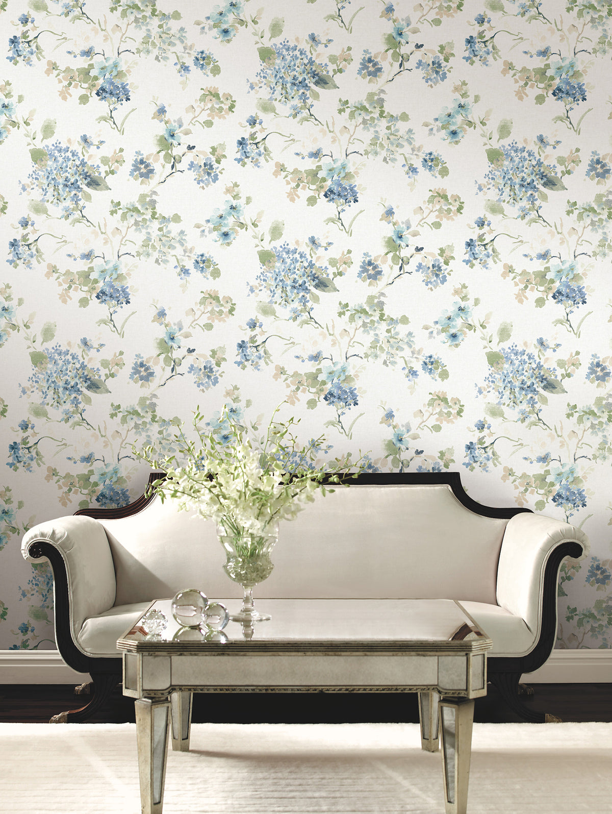 A cream sofa sits in front of a glass coffee table topped with a vase of white flowers. The background features Veiled Garden Floral Wallpaper by York Wallcoverings, adorned with blue, green, and beige flowers and leaves.