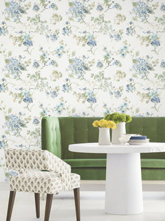 A green velvet bench and a patterned chair sit by a round white table topped with vases of yellow flowers, set against Veiled Garden Floral Wallpaper by York Wallcoverings, featuring blue and green accents.