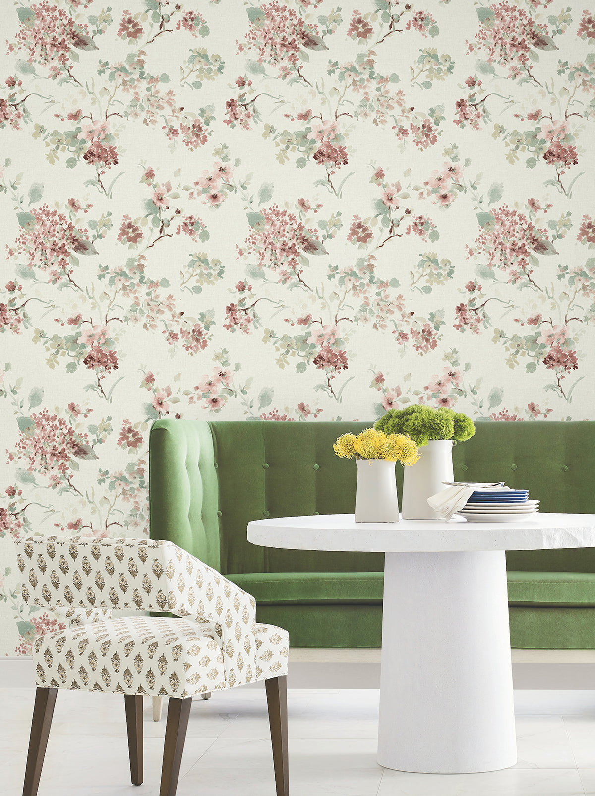A green tufted sofa and a round white table with yellow flowers sit in front of York Wallcoverings’ Veiled Garden Floral Wallpaper; an upholstered chair with a leaf pattern is nearby.