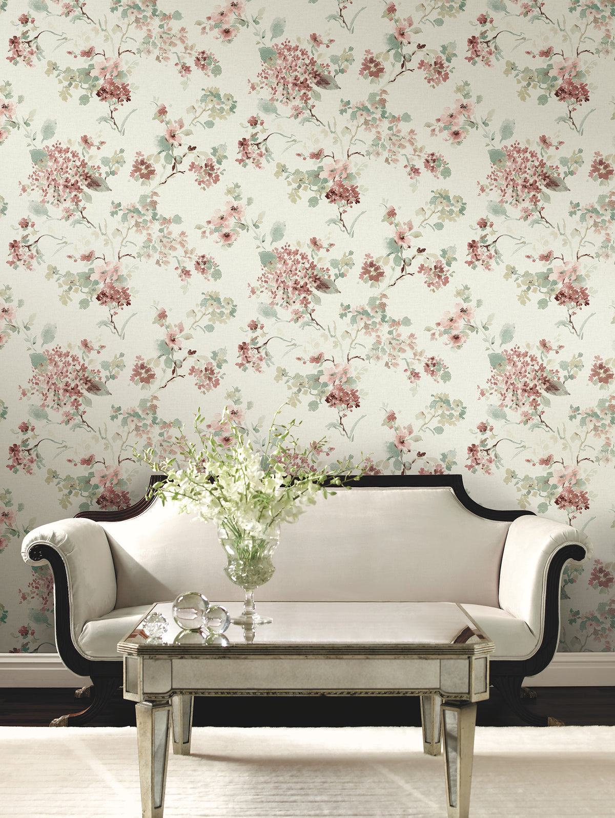An elegant white sofa with dark trim sits in front of a mirrored coffee table holding a vase of white flowers, set against a wall adorned with York Wallcoverings’ Veiled Garden Floral Wallpaper.