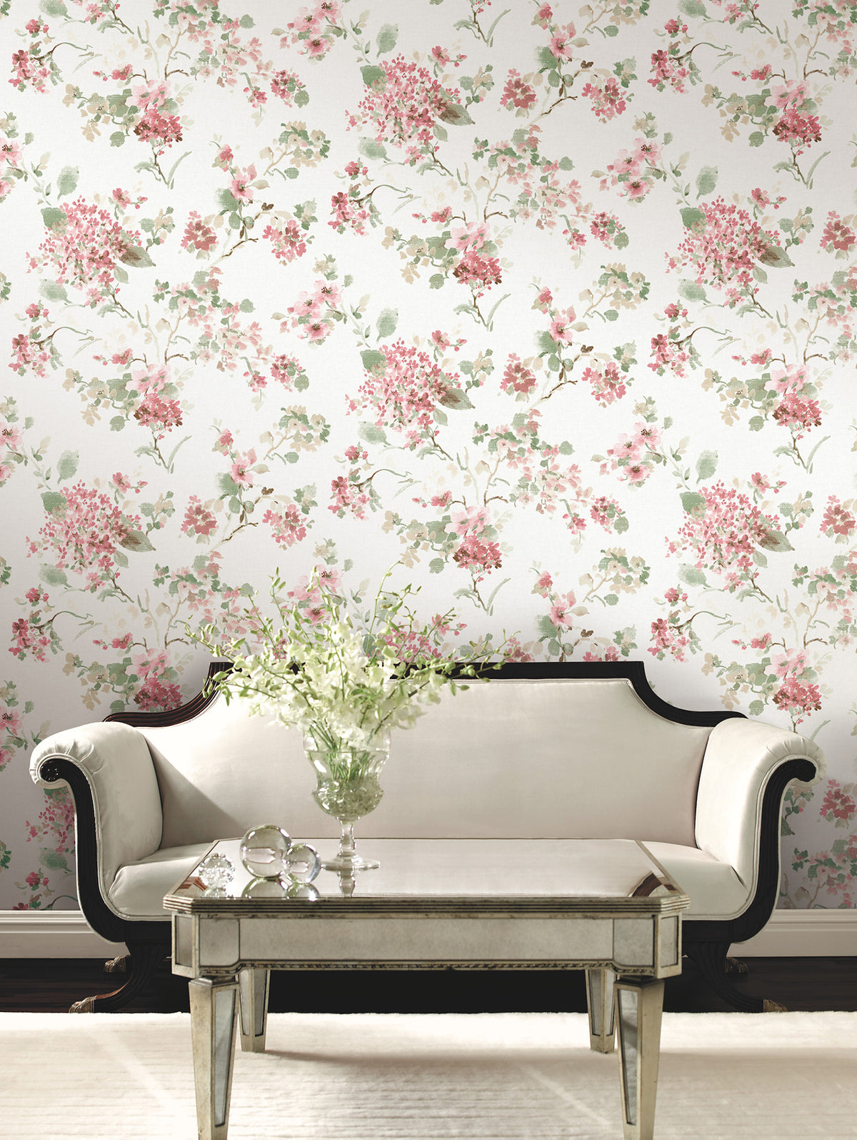 A cream sofa with dark wood trim sits before a mirrored coffee table topped with a vase of white flowers, set against a wall adorned with York Wallcoverings’ Veiled Garden Floral Wallpaper.