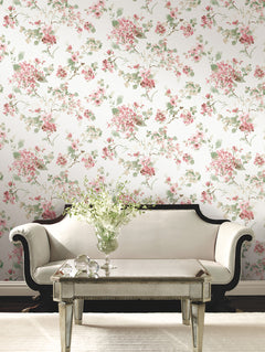 A cream sofa with dark wood trim sits before a mirrored coffee table topped with a vase of white flowers, set against a wall adorned with York Wallcoverings’ Veiled Garden Floral Wallpaper.