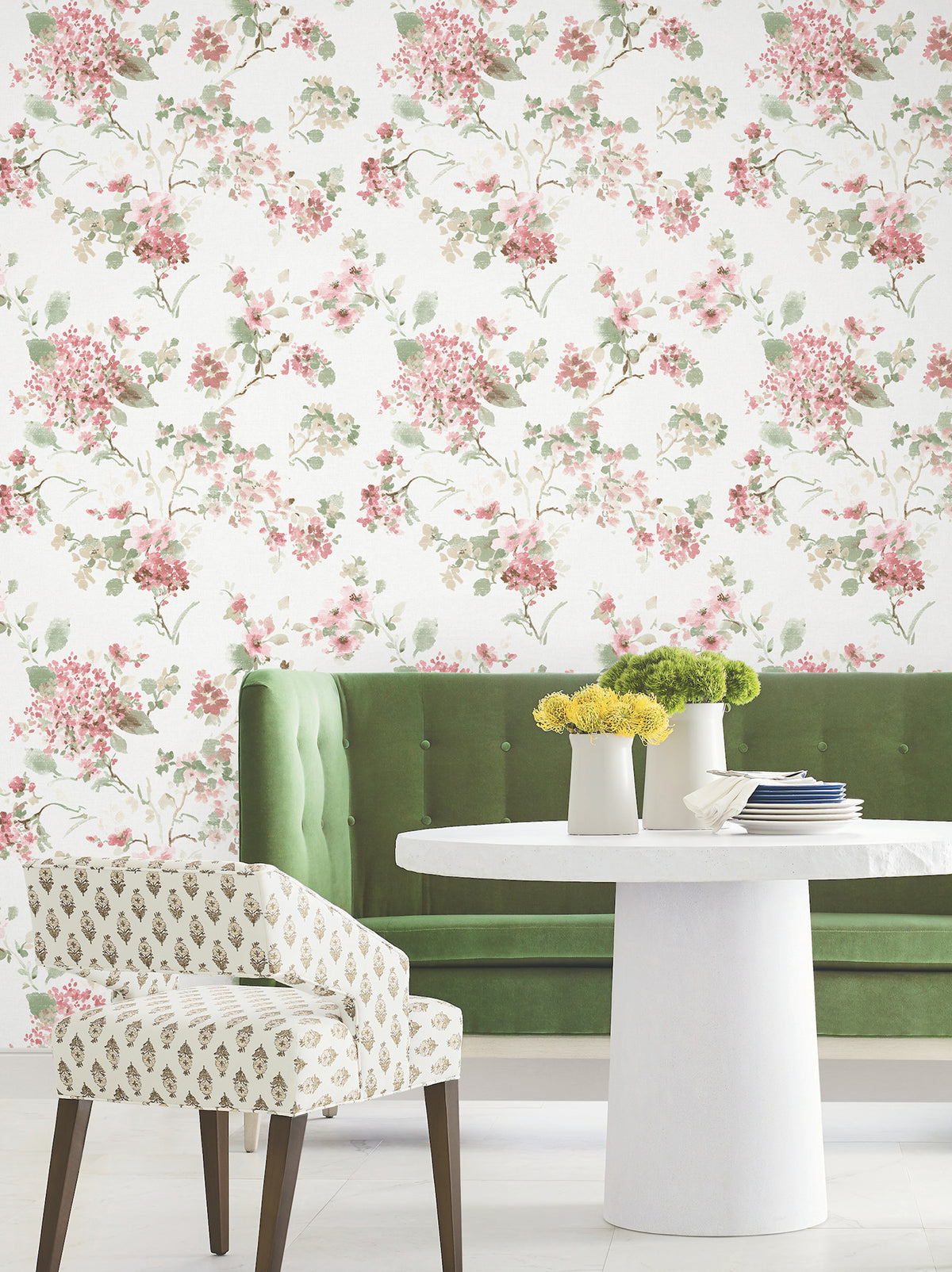 A green upholstered bench, a round white table with a vase of yellow flowers, and a patterned chair sit in front of a wall adorned with Veiled Garden Floral Wallpaper by York Wallcoverings.