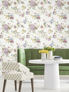A green tufted bench, round white table with yellow flowers, and patterned chair sit in front of a wall adorned with the Veiled Garden Floral Wallpaper by York Wallcoverings.