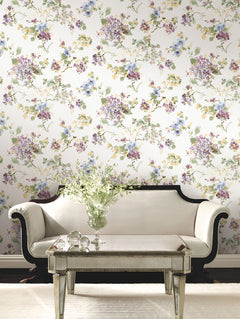A cream sofa and glass table sit in front of a wall covered in Veiled Garden Floral Wallpaper by York Wallcoverings. A vase with white flowers rests on the table, reflecting an elegant, vintage-inspired living room setting.