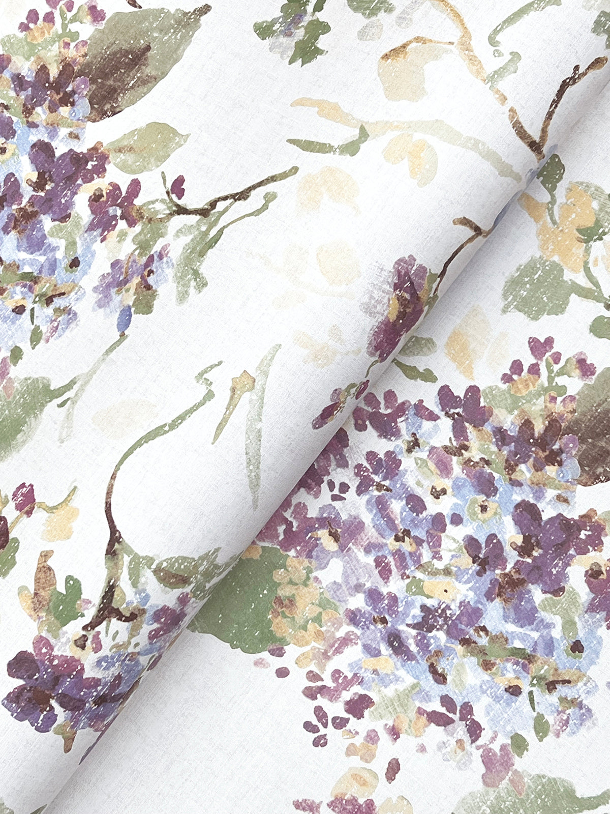 Veiled Garden Floral Wallpaper by York Wallcoverings showcases a watercolor-style floral design in purple, green, and beige on a white background, featuring leaves and scattered blossoms. The wallpaper is slightly folded to display the repeating pattern across the material.