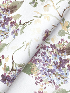 Veiled Garden Floral Wallpaper by York Wallcoverings showcases a watercolor-style floral design in purple, green, and beige on a white background, featuring leaves and scattered blossoms. The wallpaper is slightly folded to display the repeating pattern across the material.