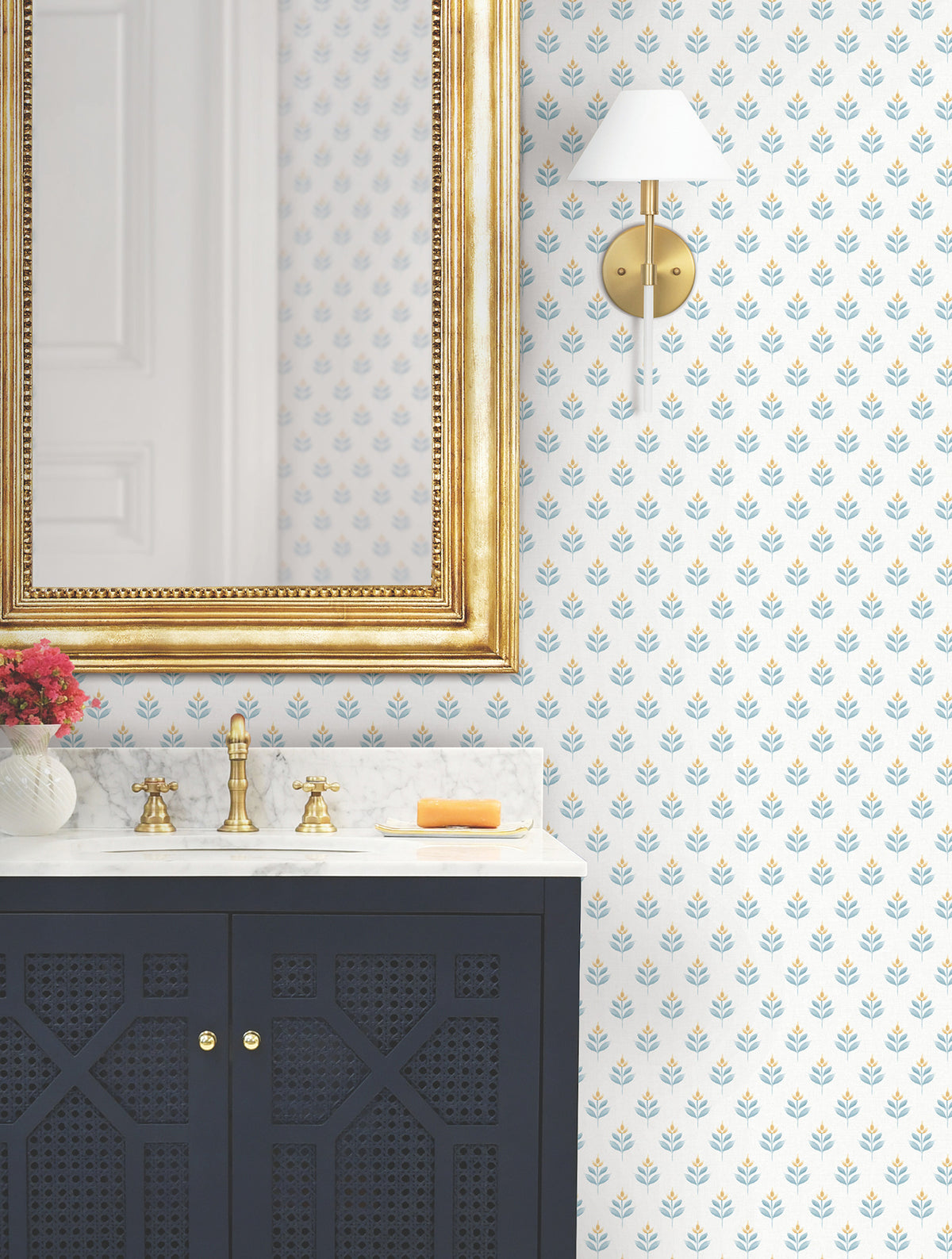 Bathroom featuring a gold-framed mirror, black vanity with marble countertop, brass faucet, soap, sconce light, and a vase with pink flowers on the counter. The walls are adorned with Serene Villa Floral Stamp Wallpaper by York Wallcoverings.