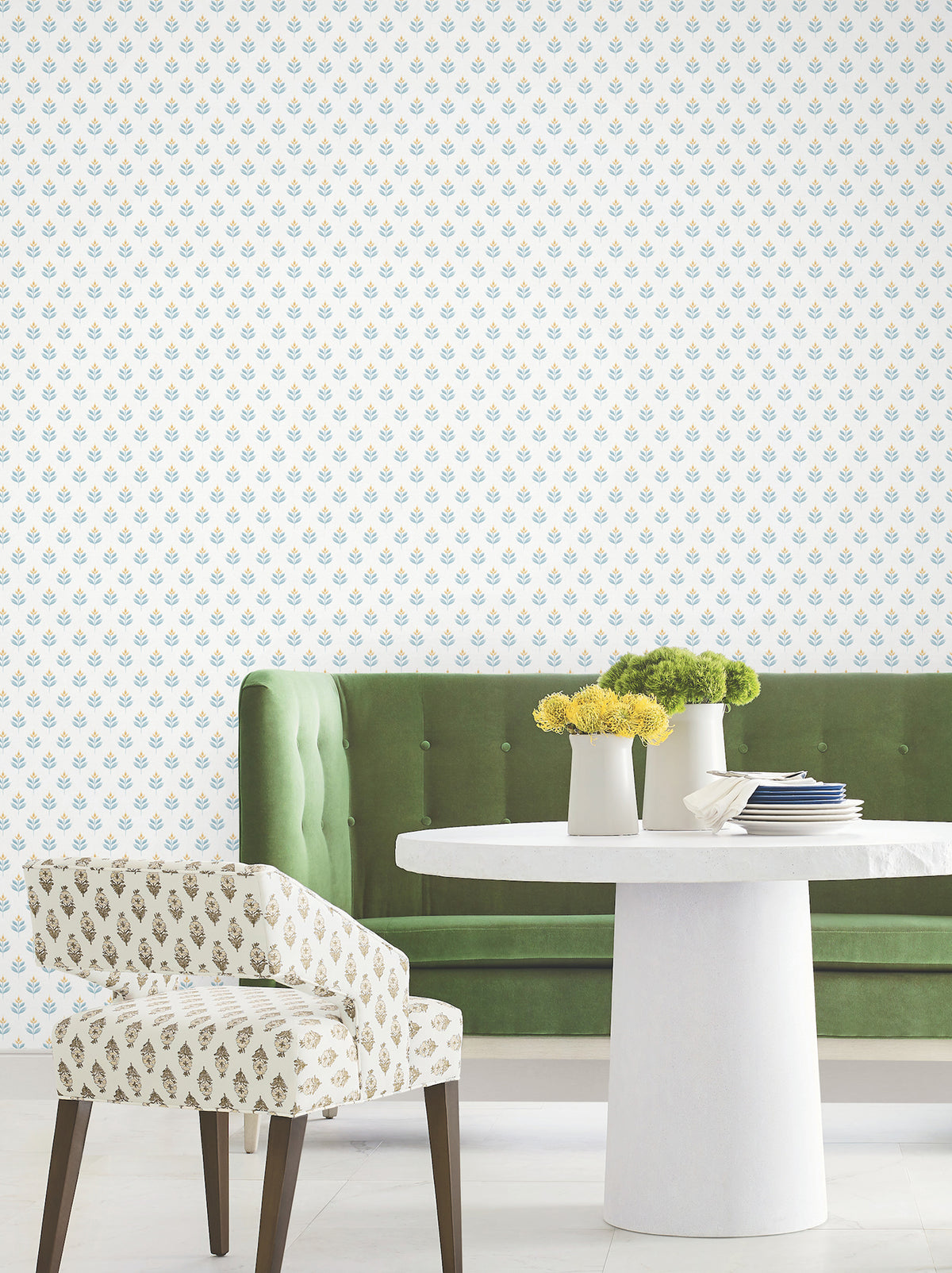A green tufted bench, a patterned chair, and a round white table with vases of yellow flowers sit in front of a white wall covered in York Wallcoverings’ Serene Villa Floral Stamp Wallpaper.
