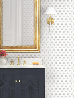 A bathroom vanity with a gold faucet, marble countertop, gold-framed mirror, wall sconce, soap bar, and a vase of flowers stands against a wall covered in Serene Villa Floral Stamp Wallpaper by York Wallcoverings—a white backdrop adorned with a delicate gray leaf pattern.