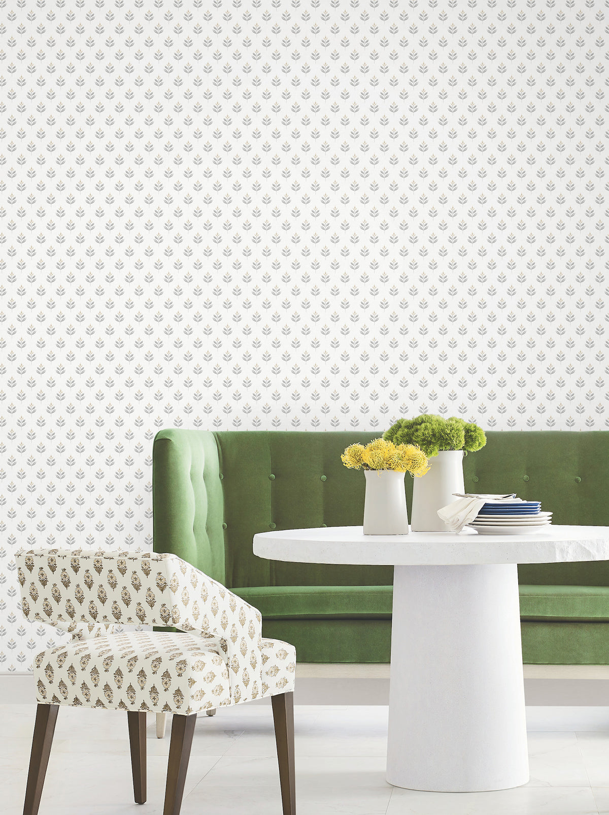 A modern dining area features a green velvet bench, a white round table, a patterned chair, and white vases filled with yellow flowers. The wall is adorned with York Wallcoverings’ Serene Villa Floral Stamp Wallpaper, showcasing a repeating small floral pattern on a white background.