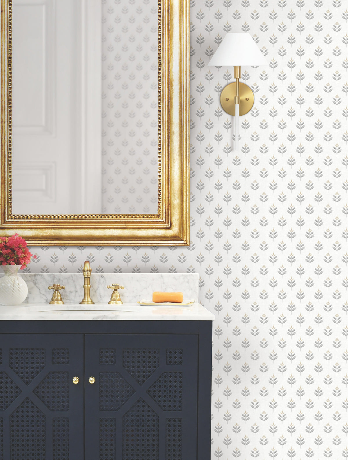 A bathroom featuring a gold-framed mirror, gold faucet, marble countertop, black vanity, a small bouquet of flowers, a bar of soap, a gold and white wall sconce, and Serene Villa Floral Stamp Wallpaper by York Wallcoverings.