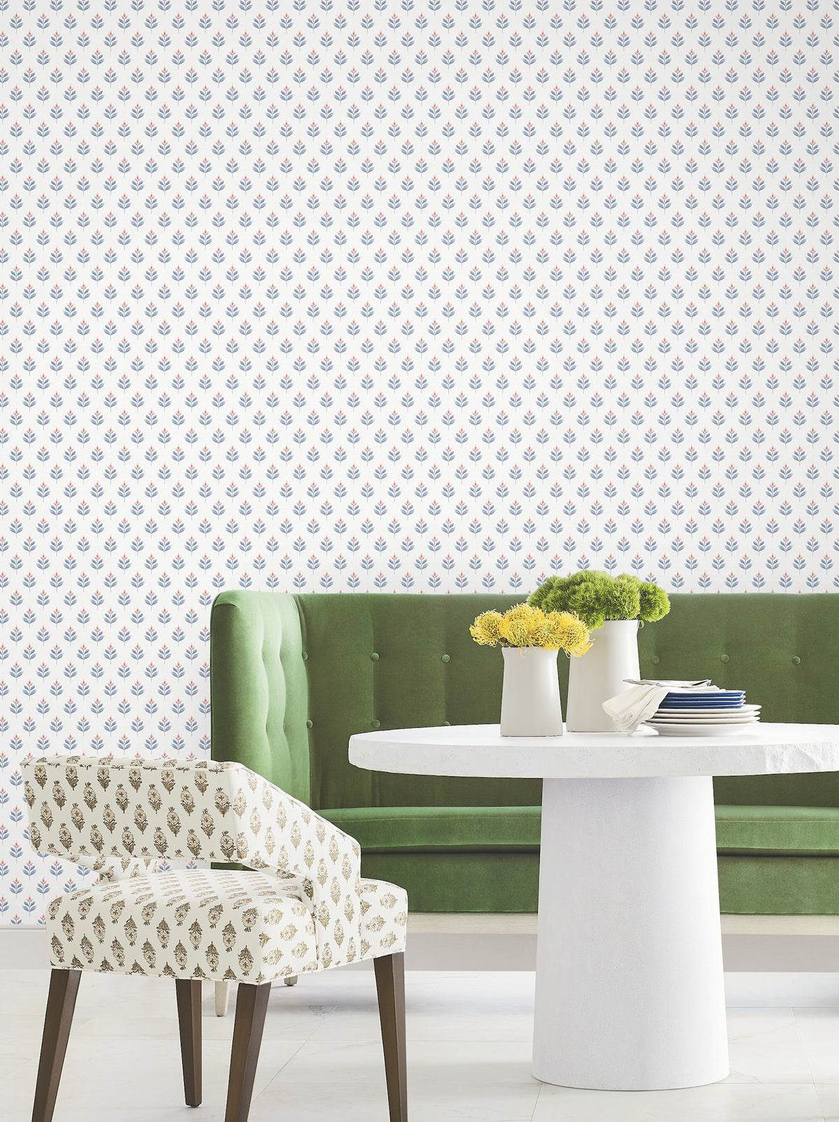 A green velvet sofa and a white round table adorned with white vases and yellow flowers are arranged in front of a wall featuring the Serene Villa Floral Stamp Wallpaper by York Wallcoverings, which displays a delicate blue and pink floral pattern. Next to the table sits a patterned chair with wooden legs.