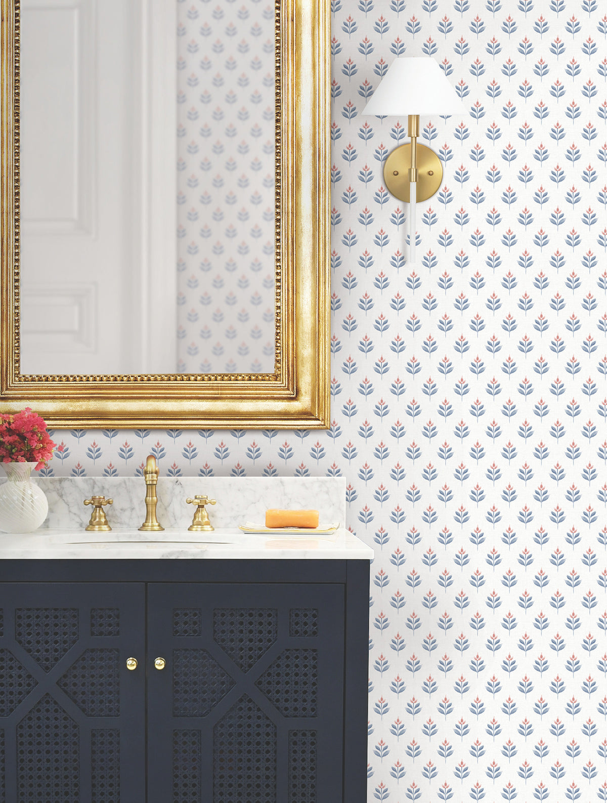 Bathroom with a gold-framed mirror, brass faucet, marble countertop, dark blue vanity, York Wallcoverings’ Serene Villa Floral Stamp Wallpaper, wall sconce, and a small vase of red flowers. Soap rests on the counter.