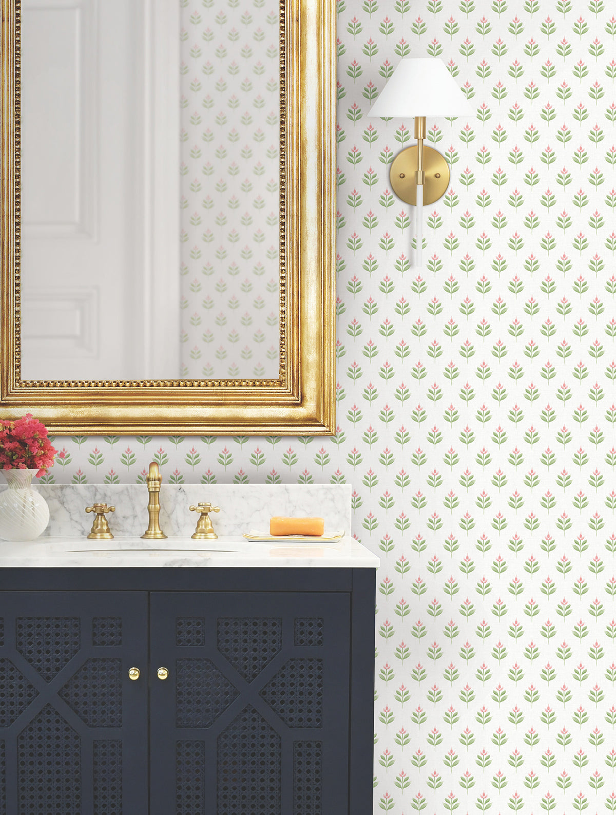 A bathroom featuring a gold-framed mirror, marble countertop, navy cabinet, brass faucet, wall sconce, orange soap, a vase with red flowers, and Serene Villa Floral Stamp Wallpaper by York Wallcoverings.