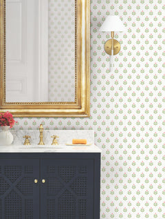 A bathroom featuring a gold-framed mirror, marble countertop, navy cabinet, brass faucet, wall sconce, orange soap, a vase with red flowers, and Serene Villa Floral Stamp Wallpaper by York Wallcoverings.