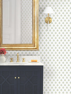 A gold-framed mirror hangs above a marble sink with a gold faucet. A navy vanity with lattice doors sits below, while a wall sconce and small bouquet of flowers are visible against York Wallcoverings’ Serene Villa Floral Stamp Wallpaper.