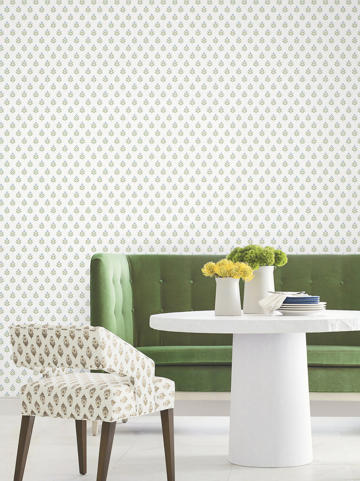 A green velvet tufted bench, a white round table with white vases holding yellow flowers, and a patterned chair are arranged against a white wall adorned with Serene Villa Floral Stamp Wallpaper by York Wallcoverings, featuring a small green and gold floral print.