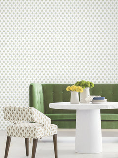 A green velvet tufted bench, a white round table with white vases holding yellow flowers, and a patterned chair are arranged against a white wall adorned with Serene Villa Floral Stamp Wallpaper by York Wallcoverings, featuring a small green and gold floral print.