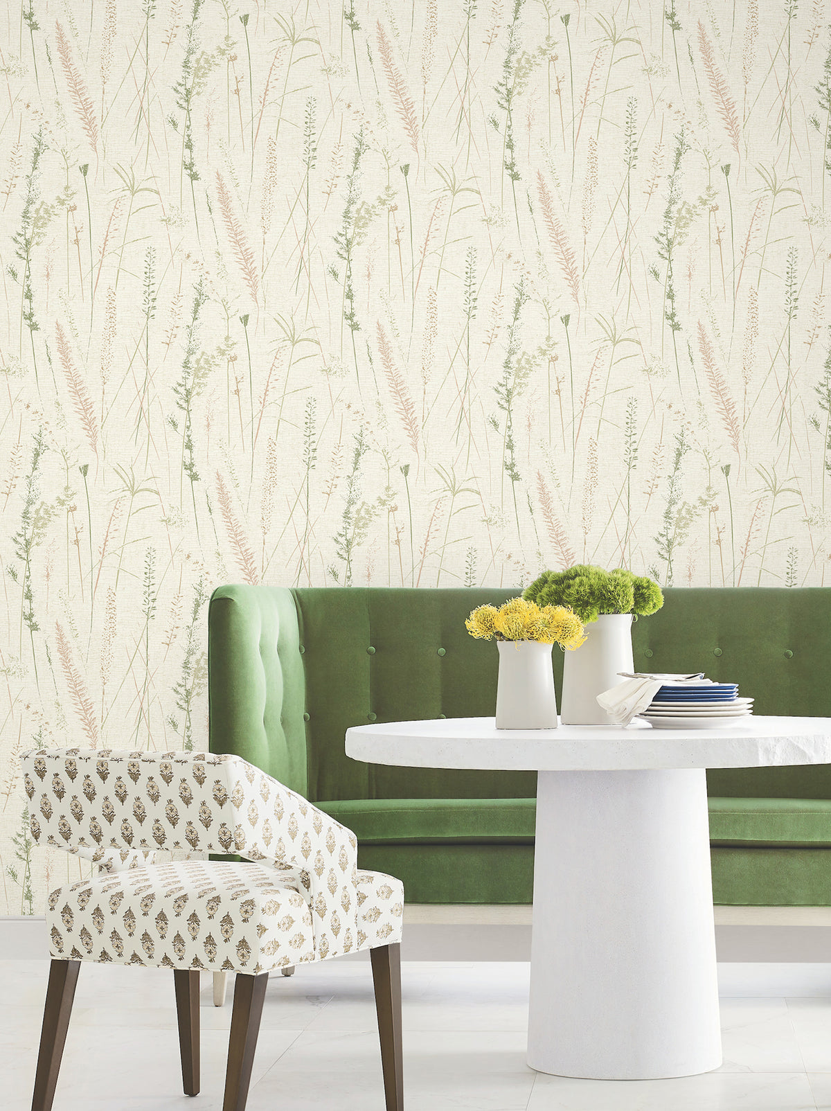 A white round table with yellow flowers, a green tufted bench, and a patterned chair sit in front of Meadow Splendor Botanical Wallpaper, featuring delicate grass and plant illustrations in soft green and beige tones.