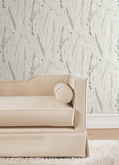 A beige sofa with a cylindrical cushion sits in front of Meadow Splendor Botanical Wallpaper, which features a delicate pattern of grasses and plants in light tones.