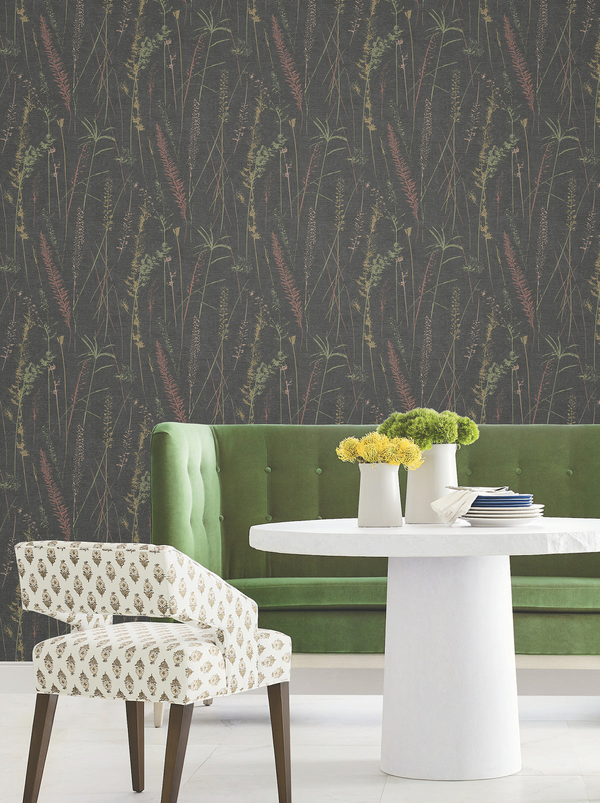 A modern dining area with a green velvet tufted bench, a white round table topped with yellow flowers, a patterned chair, and dark Meadow Splendor Botanical Wallpaper.