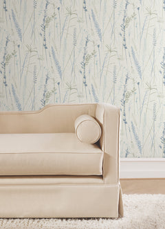 A beige upholstered sofa with a round bolster pillow sits on a wooden floor in front of Meadow Splendor Botanical Wallpaper featuring a light botanical pattern with blue and green grasses.