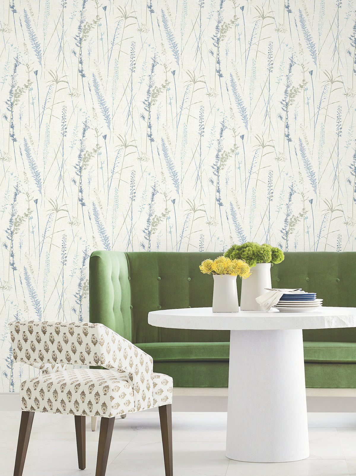 A green velvet bench, a white round table with yellow flowers, and a patterned chair sit in front of Meadow Splendor Botanical Wallpaper featuring light blue and green botanical designs.