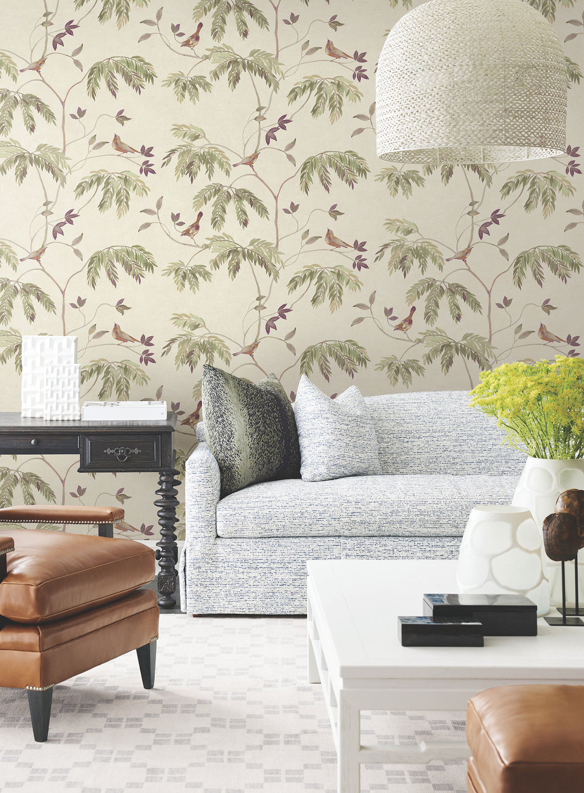 A stylish living room featuring the Cillian Botanical Bird Trail Wallpaper by York Wallcoverings, a light blue sofa, leather ottoman, white coffee table, black side table, vases with yellow flowers, and a textured hanging lamp. The space feels bright and inviting.