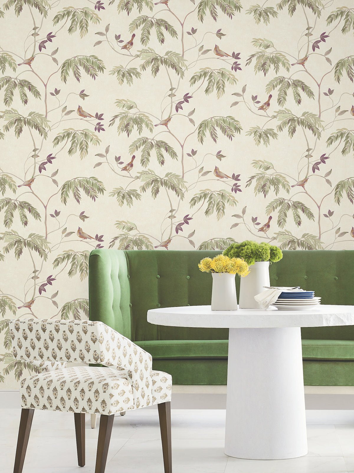 A green tufted sofa, patterned chair, and round white table topped with yellow and green floral arrangements sit in front of York Wallcoverings’ Cillian Botanical Bird Trail Wallpaper, featuring leafy branches and small birds on a cream background.