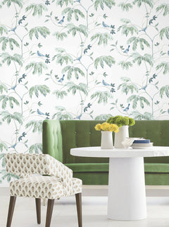 A green velvet bench and patterned chair sit by a round white table adorned with vases of yellow flowers, set against a backdrop of York Wallcoverings’ Cillian Botanical Bird Trail Wallpaper.