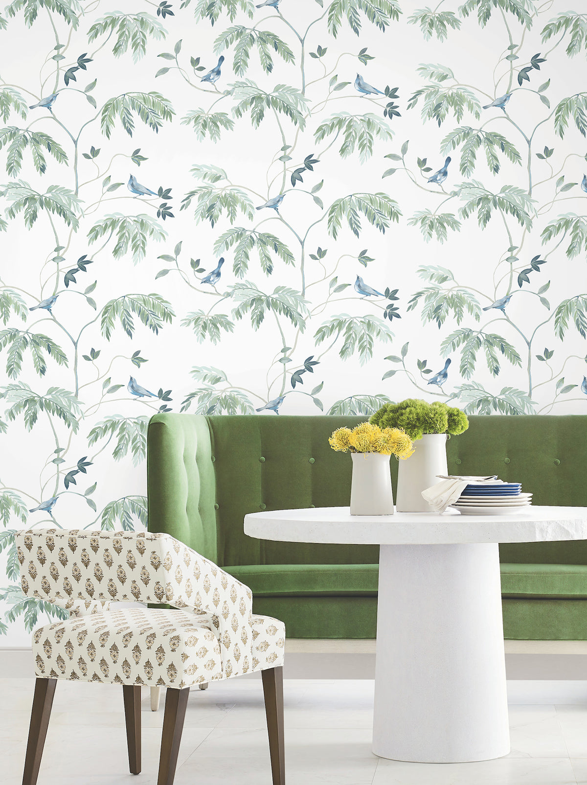 A white table with yellow flowers, a green velvet bench, and a patterned chair sit in front of a wall adorned with Cillian Botanical Bird Trail Wallpaper by York Wallcoverings.