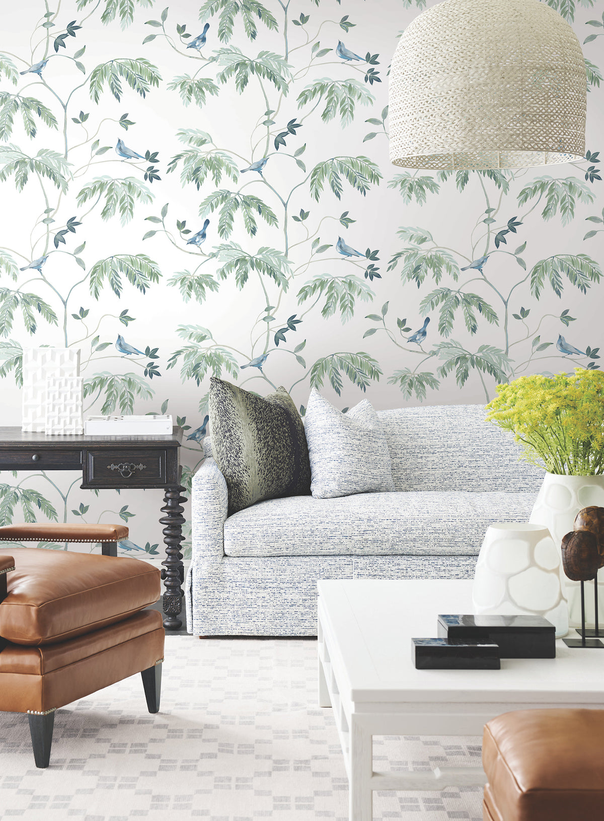 A bright living room with York Wallcoverings’ Cillian Botanical Bird Trail Wallpaper, a light sofa with pillows, a black side table, a white coffee table, a brown leather armchair, a vase of yellow flowers, and a woven pendant lamp.