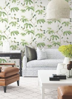 A stylish living room with York Wallcoverings’ Cillian Botanical Bird Trail Wallpaper, a light blue sofa, leather armchair, black side table, white coffee table, yellow flowers, and a large textured hanging lamp.