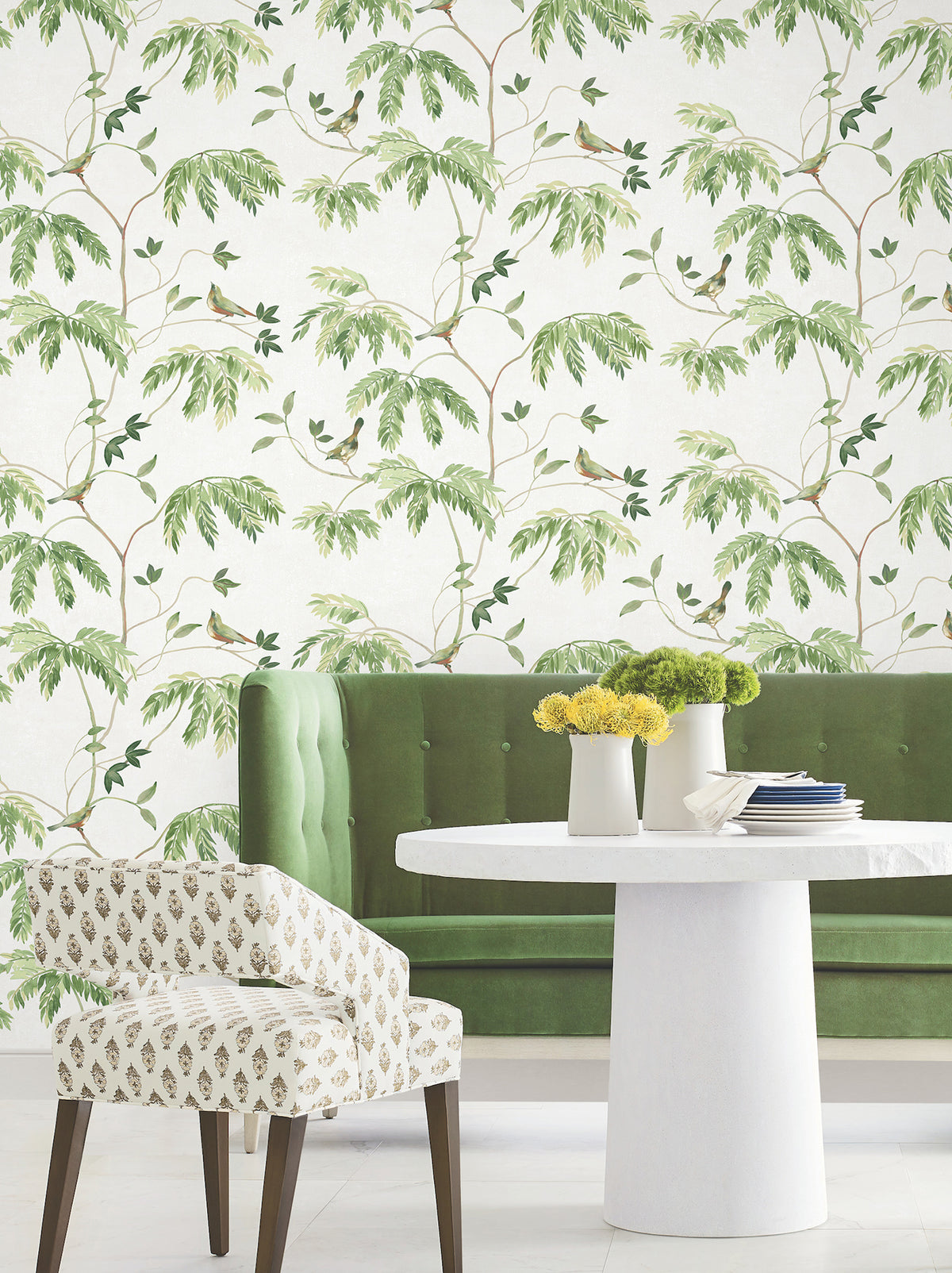 Green velvet bench and patterned chair surround a white round table with yellow flowers, set against York Wallcoverings’ Cillian Botanical Bird Trail Wallpaper.