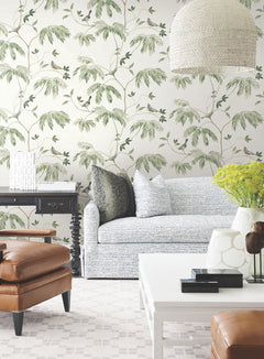 A bright living room featuring the Cillian Botanical Bird Trail Wallpaper by York Wallcoverings, a light blue sofa, brown leather chair, white tables, green and black pillows, yellow flowers, and a woven lampshade hanging from the ceiling.