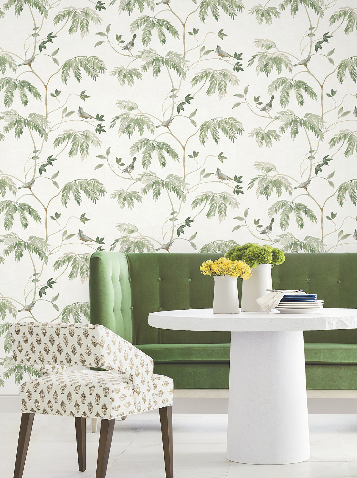 A green velvet sofa, a white round table adorned with yellow flowers in vases, and a patterned chair sit in front of the Cillian Botanical Bird Trail Wallpaper by York Wallcoverings, creating a light and airy atmosphere.