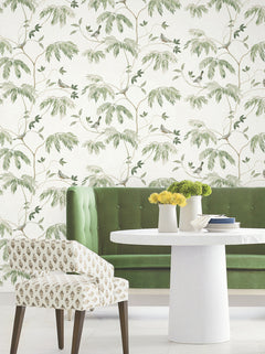 A green velvet sofa, a white round table adorned with yellow flowers in vases, and a patterned chair sit in front of the Cillian Botanical Bird Trail Wallpaper by York Wallcoverings, creating a light and airy atmosphere.