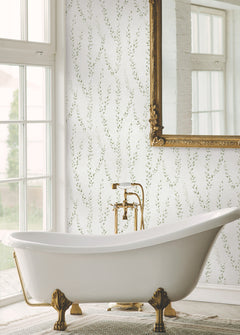 An elegant white clawfoot bathtub with gold feet and fixtures is positioned before a spacious window. The wall behind features Alpenrose Vine Wallpaper by York Wallcoverings, showcasing a delicate leafy design, while a grand ornate gold-framed mirror hangs above. Sunlight pours in, filling the bathroom with a calm and refined ambiance.