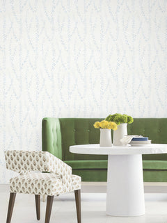 A green tufted sofa, a white round table with vases of yellow flowers, and a patterned chair are set against a white wall adorned with York Wallcoverings’ Alpenrose Vine Wallpaper.