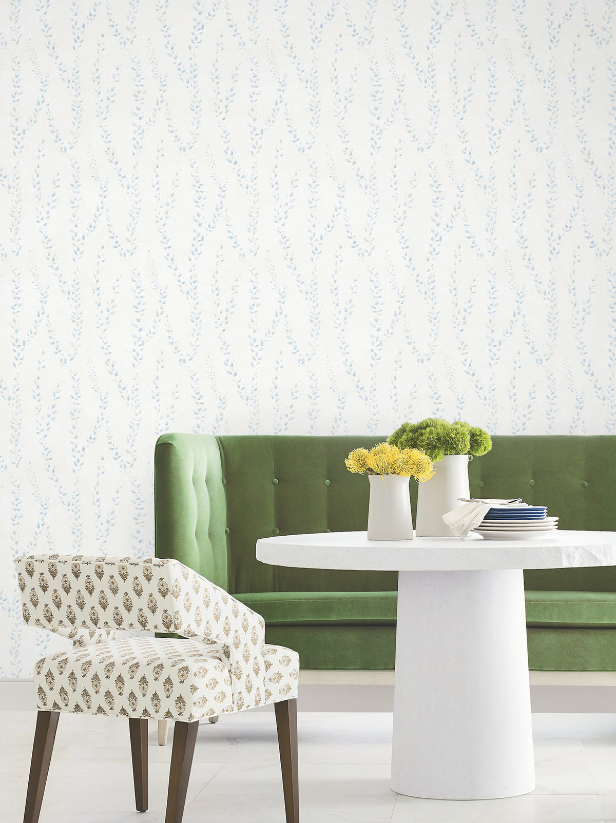A green tufted sofa and a patterned chair sit by a round white table with three vases of yellow flowers, set against York Wallcoverings’ Alpenrose Vine Wallpaper.
