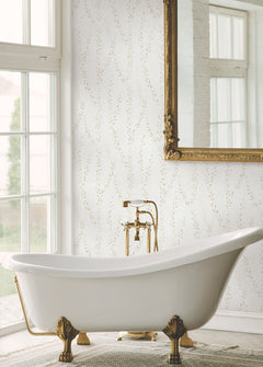 A white clawfoot bathtub with gold fixtures sits by a large window. The wall is adorned with Alpenrose Vine Wallpaper from York Wallcoverings, featuring a delicate, light pattern, and a large ornate gold-framed mirror hangs above the tub.
