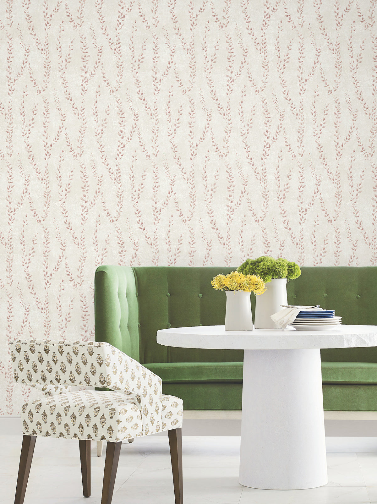 A green tufted sofa, a round white table adorned with yellow flowers in vases, and a patterned chair are arranged against walls covered in York Wallcoverings’ Alpenrose Vine Wallpaper.