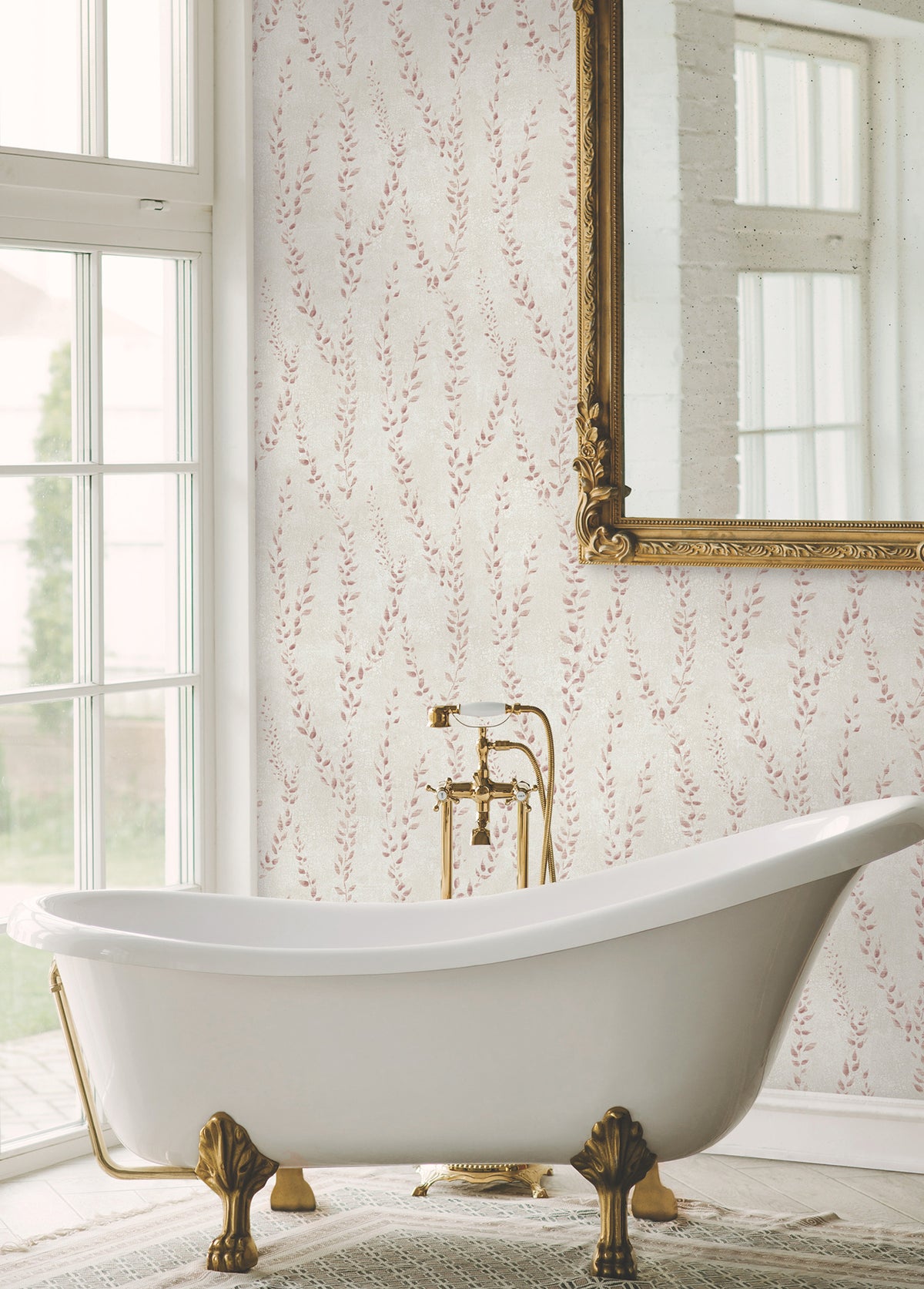 An elegant white clawfoot bathtub with gold fixtures sits in front of a large window. The wall is adorned with Alpenrose Vine Wallpaper by York Wallcoverings, featuring a subtle pink leaf pattern, and a large ornate mirror hangs above the tub.