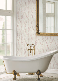 An elegant white clawfoot bathtub with gold fixtures sits in front of a large window. The wall is adorned with Alpenrose Vine Wallpaper by York Wallcoverings, featuring a subtle pink leaf pattern, and a large ornate mirror hangs above the tub.