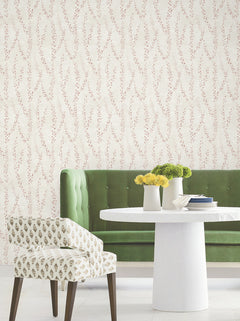 A green tufted sofa, a white round table with white vases holding yellow flowers, and a patterned upholstered chair are arranged in front of beige Alpenrose Vine Wallpaper by York Wallcoverings, featuring a delicate red vine design.