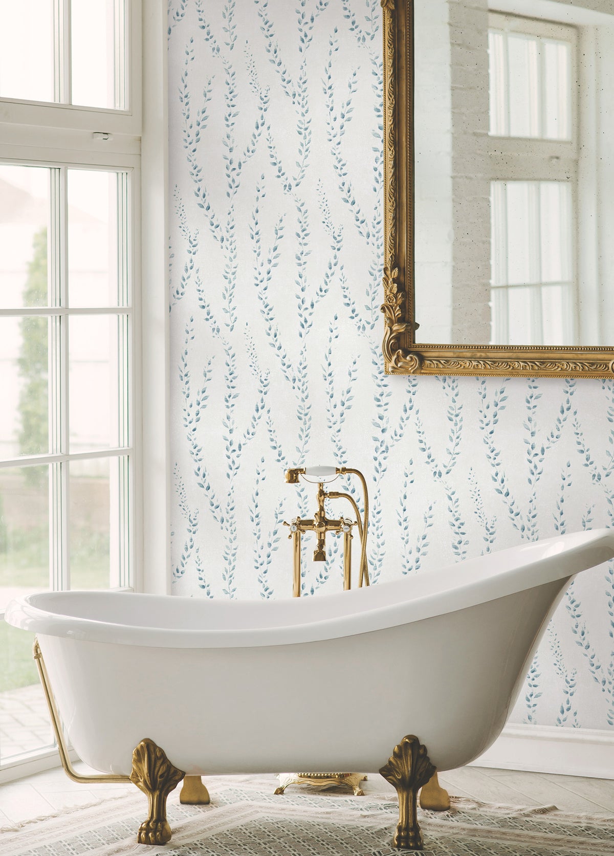 An elegant white clawfoot bathtub with gold fixtures is placed by a large window. Behind it, the wall features Alpenrose Vine Wallpaper by York Wallcoverings in a blue leaf pattern, and a large ornate gold-framed mirror reflects the view outside.
