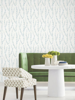 A green tufted sofa, round white table with small vases of yellow flowers, and a patterned chair sit in front of a white wall adorned with York Wallcoverings’ Alpenrose Vine Wallpaper.