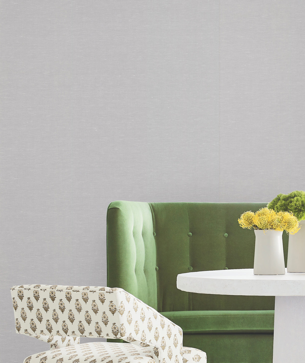 A green velvet sofa and a patterned chair are arranged beside a white round table with two small potted yellow flowers, set against walls adorned with York Wallcoverings’ Hempfield Weave Loomed Wallpaper.